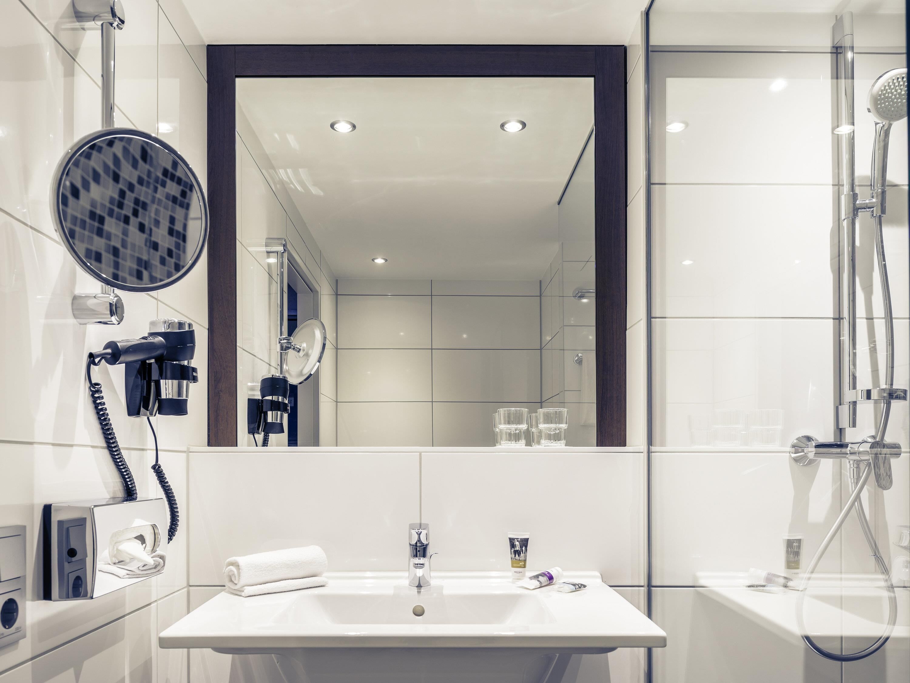 A modern bathroom with a large mirror, a hairdryer, a towel, a sink, and a shower.