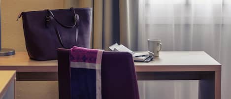 Quarto de hotel com uma escrivaninha, uma cadeira, uma bolsa grande, um caderno, uma caneta e uma xícara.