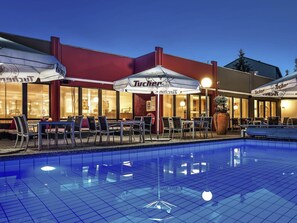 A poolside area with outdoor seating and umbrellas, a building with large windows, and a sign reading 'Tucher'.