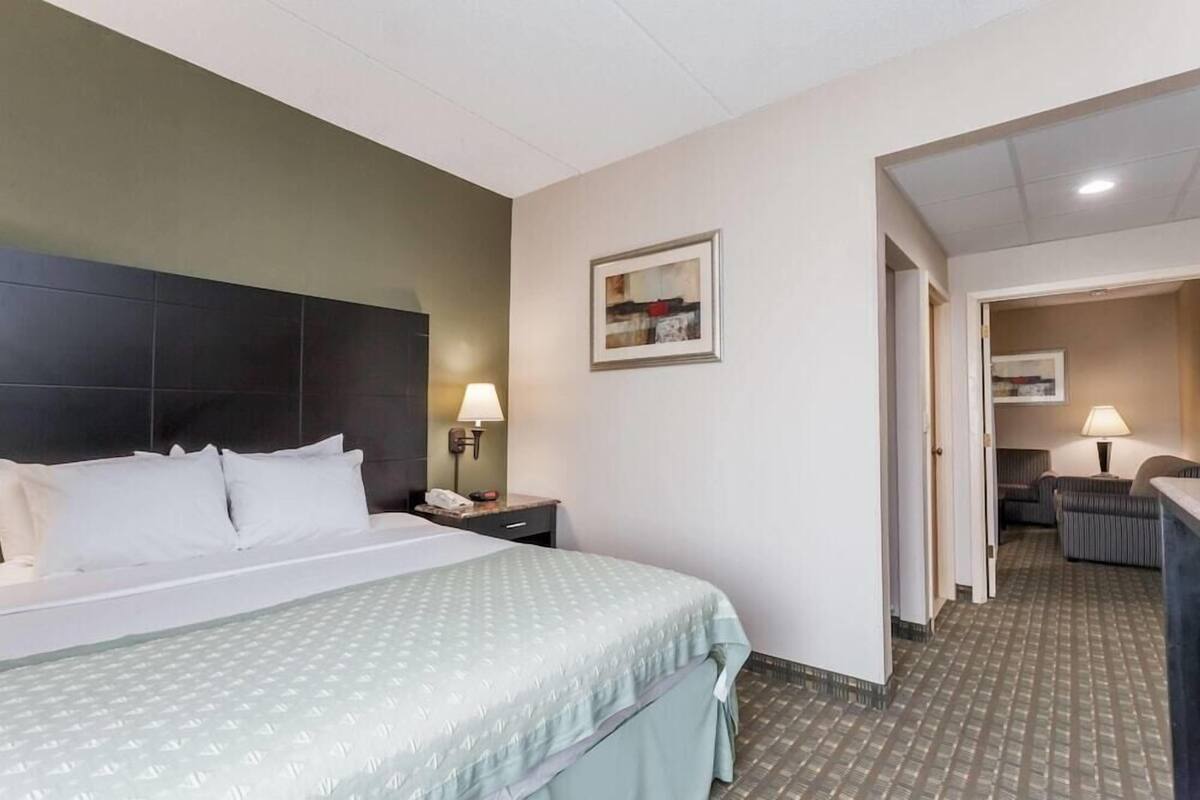 A hotel room with a bed, a nightstand, a lamp, a framed picture on the wall, and a hallway leading to a seating area.