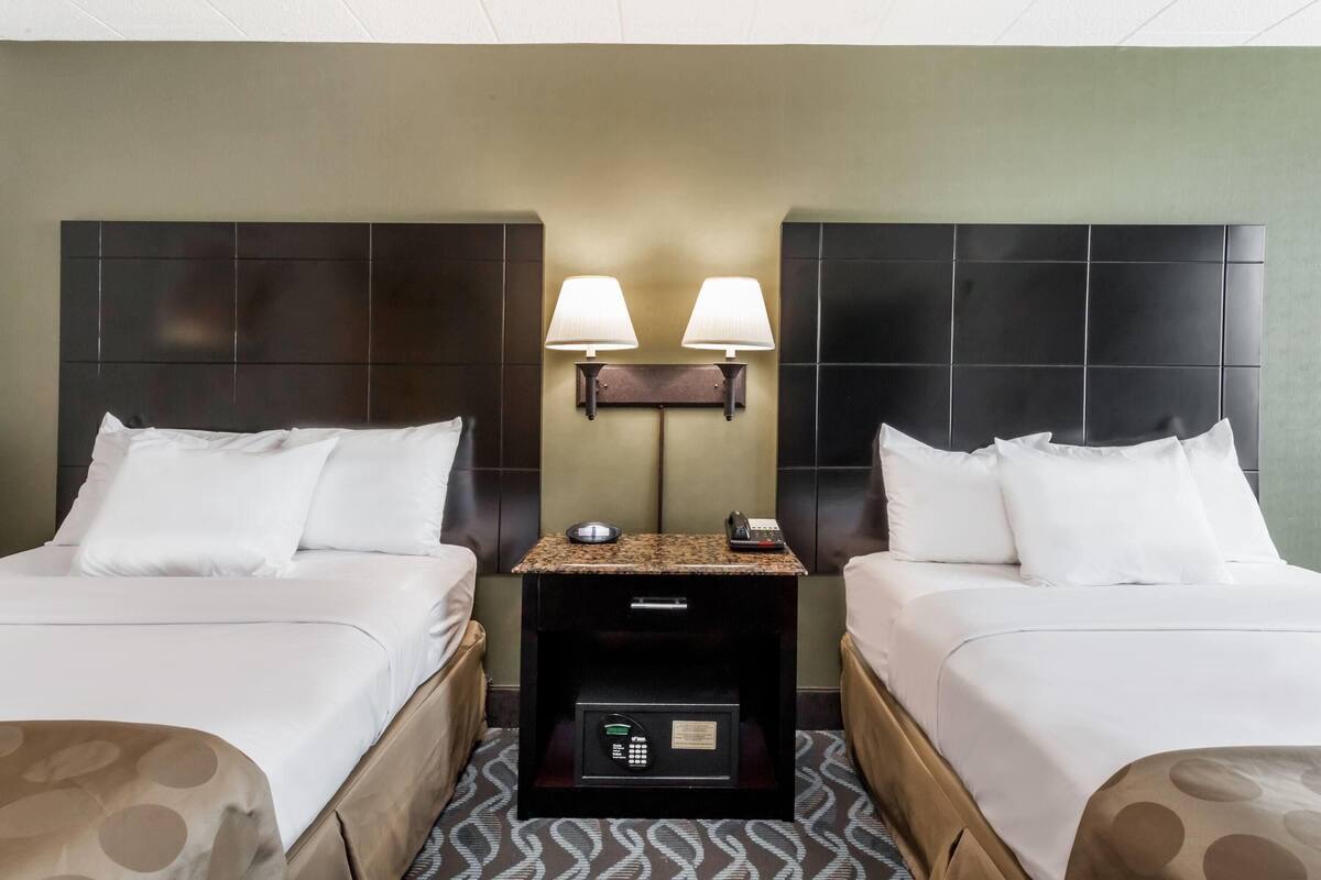 A hotel room with two beds, a nightstand with a lamp, a safe, and a wall-mounted phone.