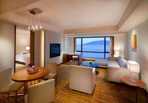 Una camera d'hotel con un'ampia finestra vista mare, un'area pranzo con tavolo e sedie, un divano e un piccolo tavolino con lampada.