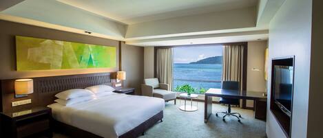 A hotel room with a large bed, a desk, a chair, and a TV. There is a window with a view of the sea.