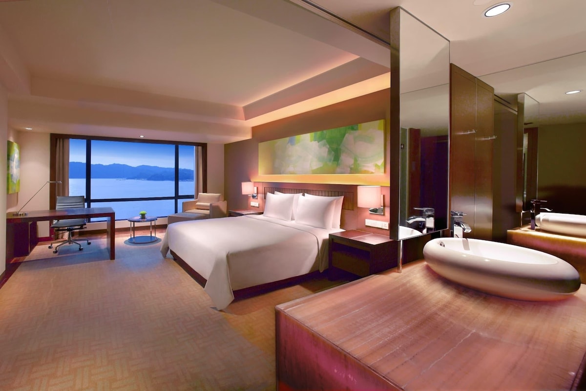 A hotel room with a bed, desk, chair, and a large window offering a view of the sea.