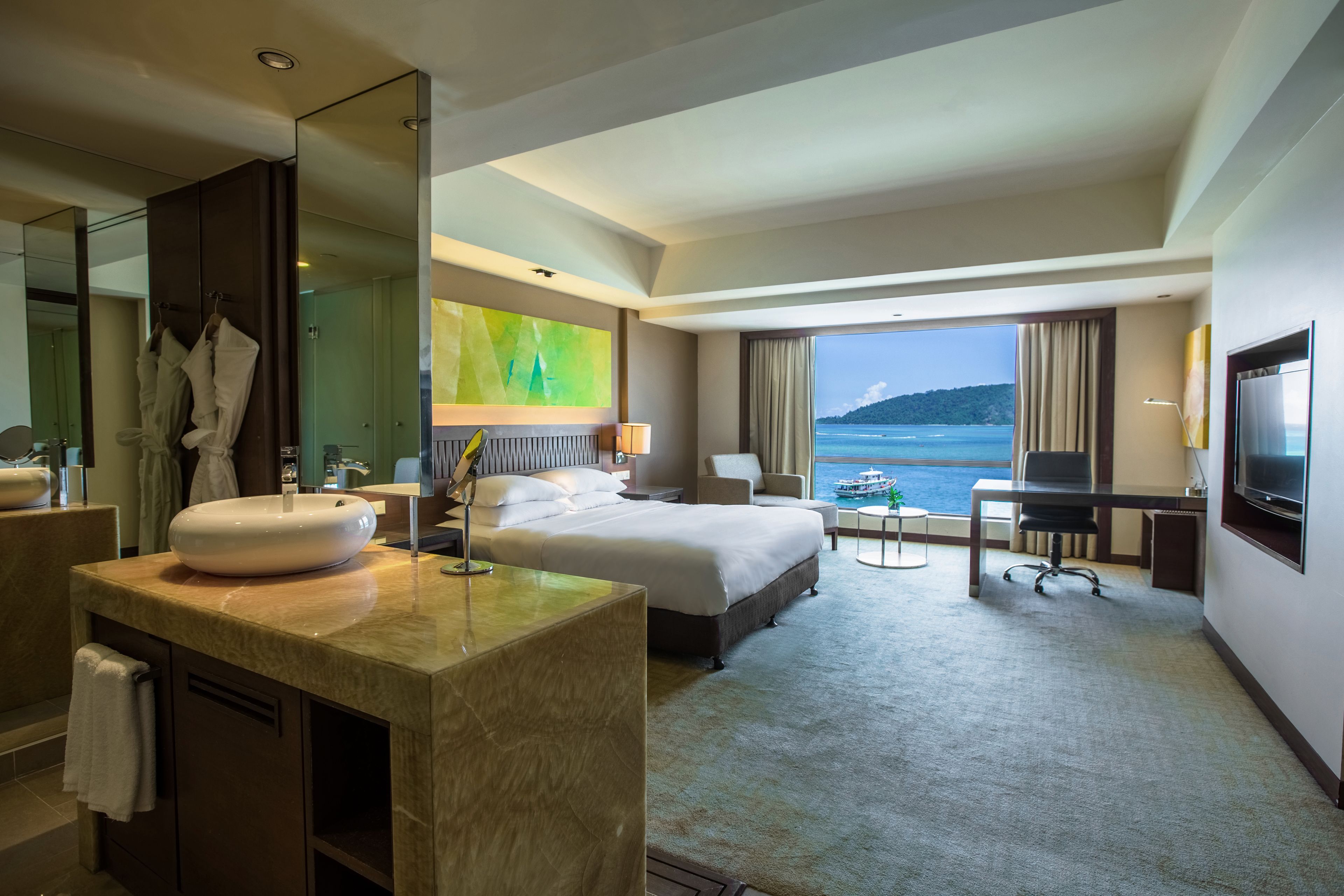 Ein Hotelzimmer mit einem großen Bett, einem Schreibtisch, einem Fernseher, einem Badezimmer mit Waschbecken und Spiegel sowie Blick aufs Meer.