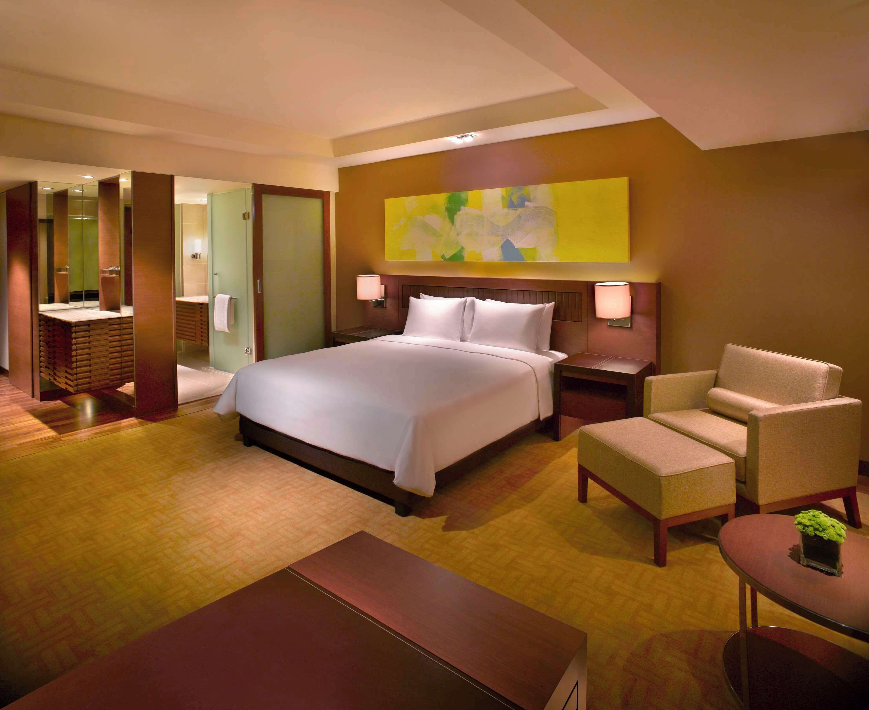 Una habitación de hotel con una cama grande, una zona de estar con una silla y una mesa, un escritorio pequeño y un cuadro abstracto de grandes dimensiones en la pared.