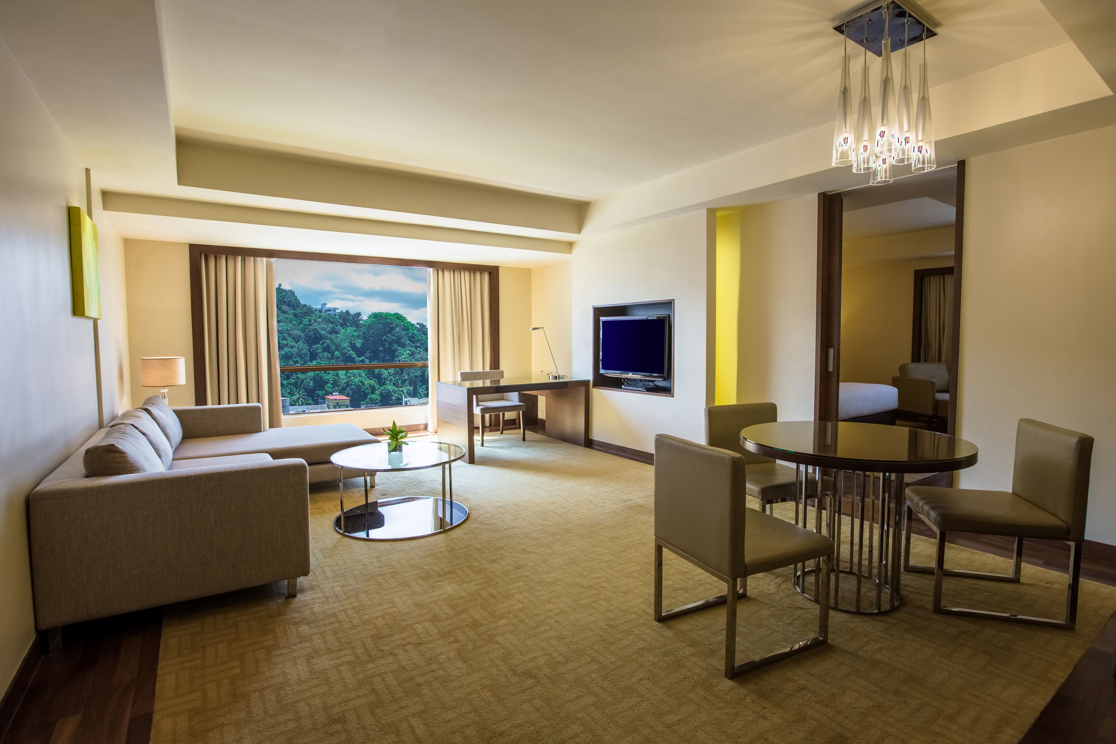 Een moderne hotelkamer met een bank, een eettafel, stoelen, een televisie en een groot raam met uitzicht op groen.