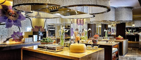 A luxurious hotel lobby with a central island featuring a cheese wheel, fruit bowls, and a coffee station.