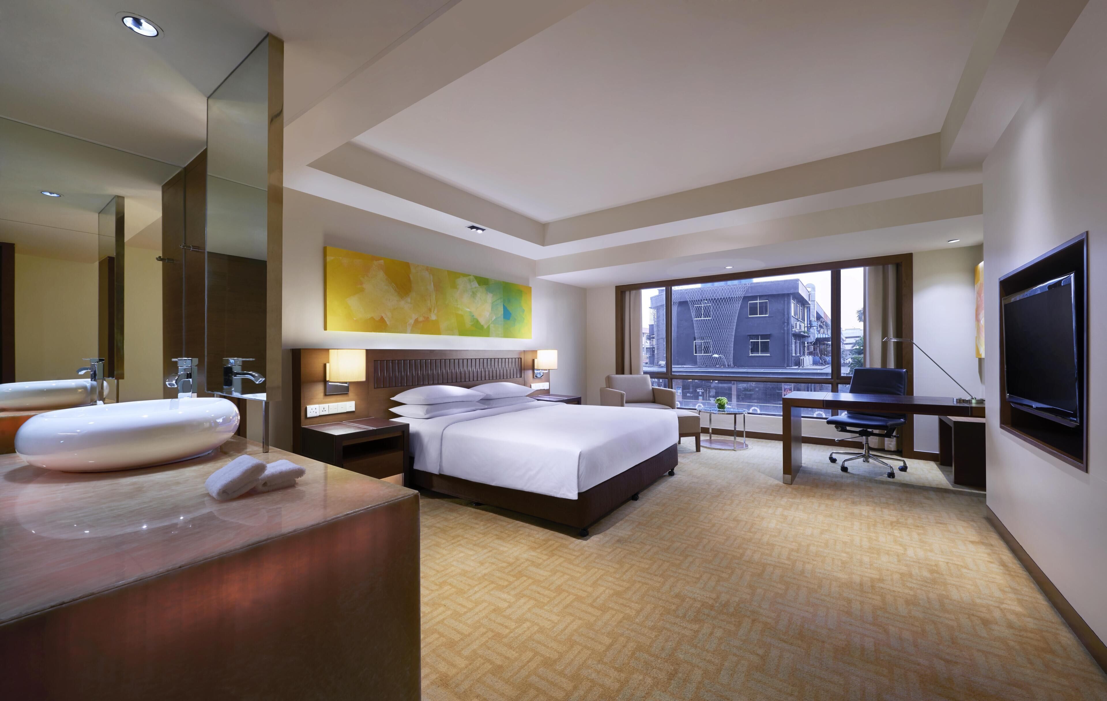 A hotel room with a large bed, a desk, a TV, a bathroom with a large mirror, and a city view.