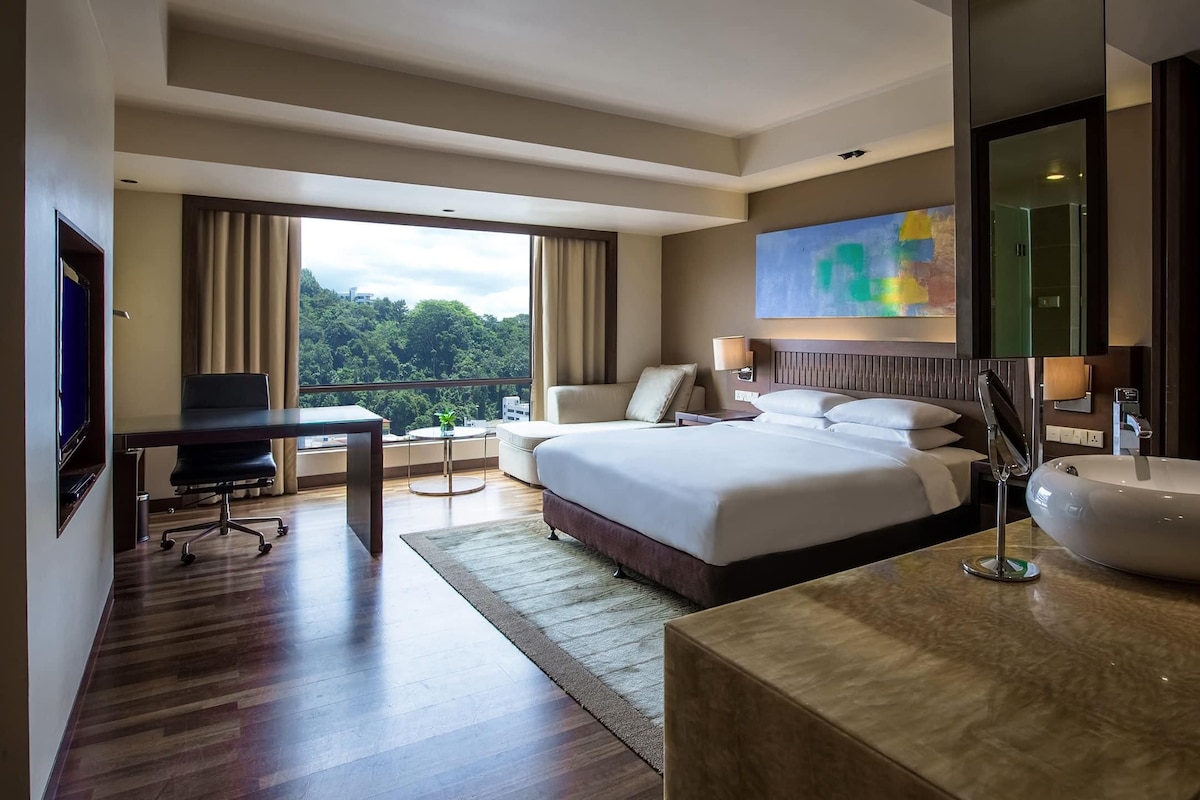 A hotel room with a large bed, a desk, a chair, a large window, and a bathroom with a sink and mirror.