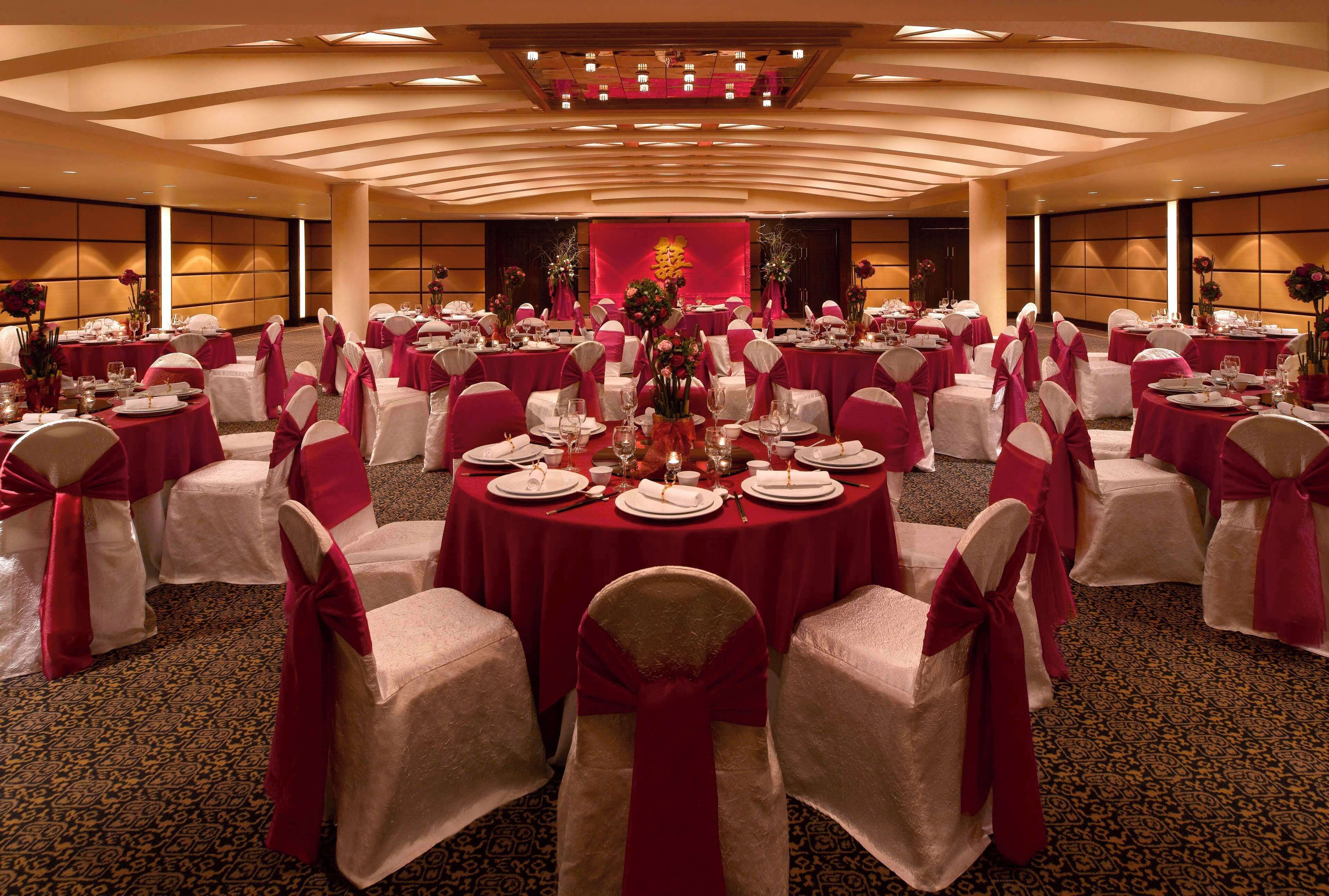 A banquet hall with round tables, white chairs with red sashes, and a red backdrop with a golden emblem.