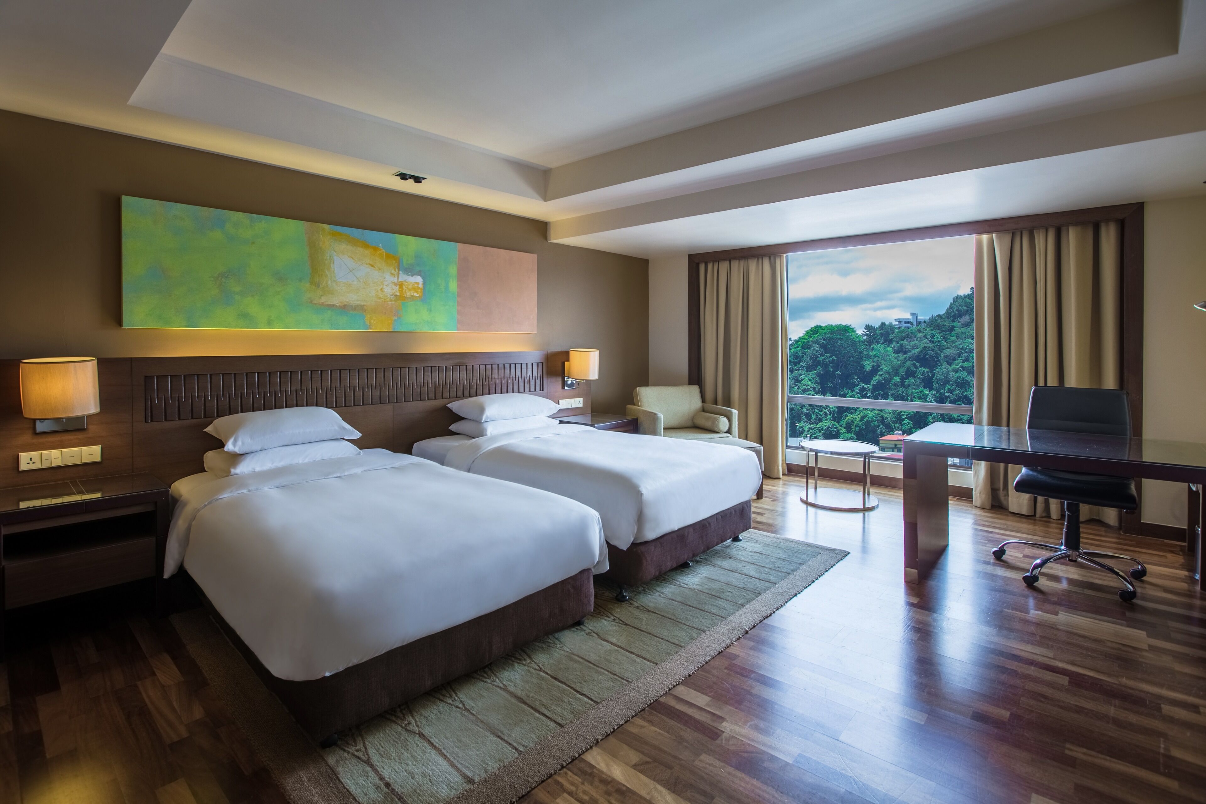 Habitación de hotel con dos camas, un escritorio, una silla y un ventanal con vistas a zonas verdes.