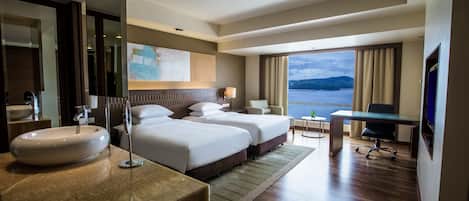 Habitación de hotel con dos camas, un escritorio, televisión, ventana grande con vista al mar y baño con lavamanos y espejo.