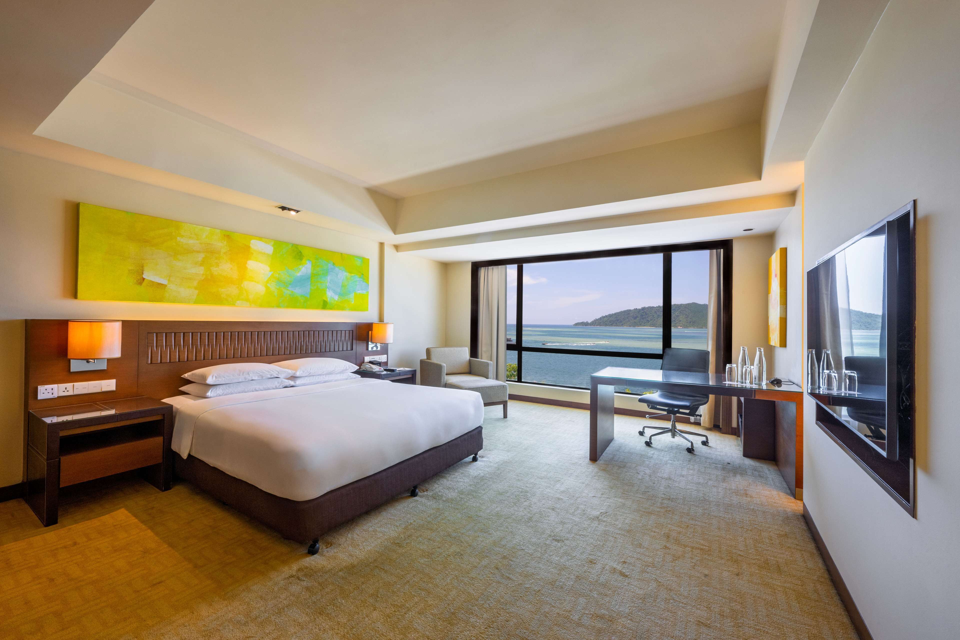 A hotel room with a large bed, a desk, a chair, a TV, and a view of the sea.