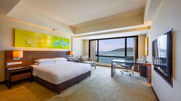 A hotel room with a large bed, a desk, a chair, a TV, and a view of the sea.