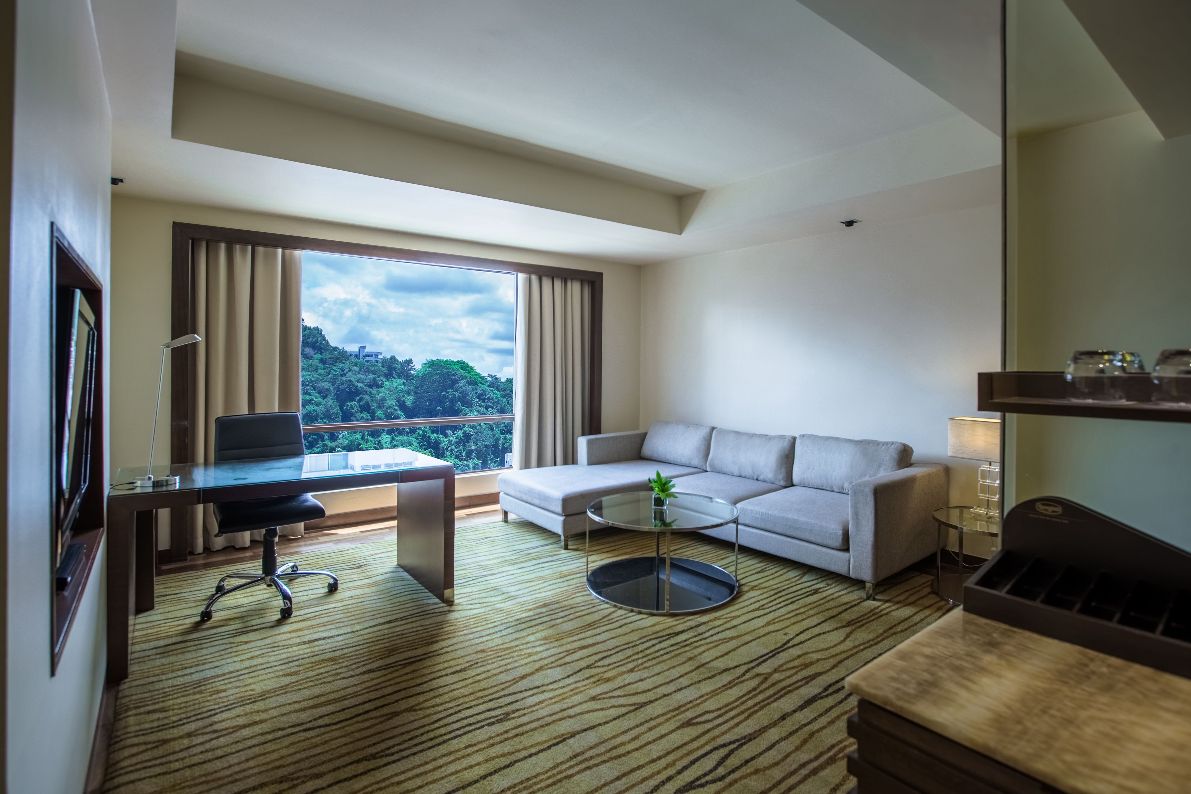 A modern hotel room with a large window, a sofa, a desk with a chair, and a small coffee table.