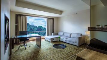 A modern hotel room with a large window, a sofa, a desk with a chair, and a small coffee table.