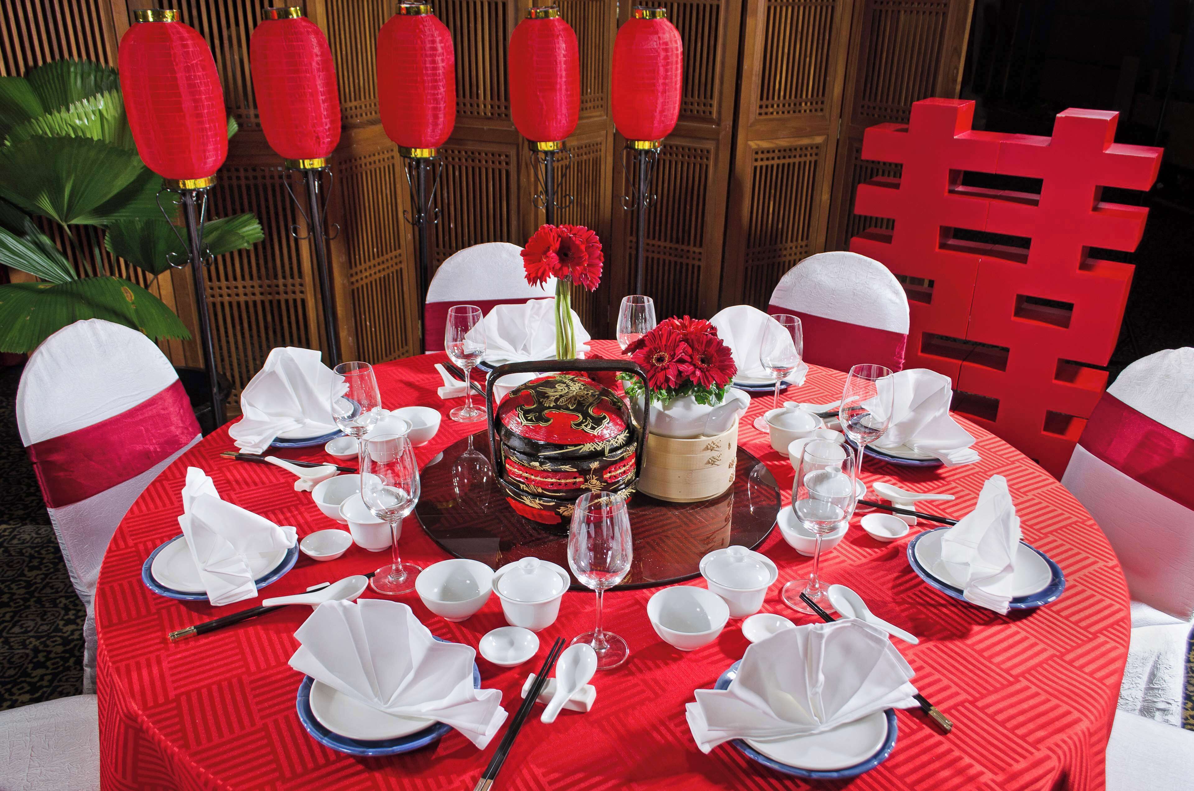 A round dining table set with white and blue plates, bowls, and cutlery, surrounded by red chairs with white sashes.