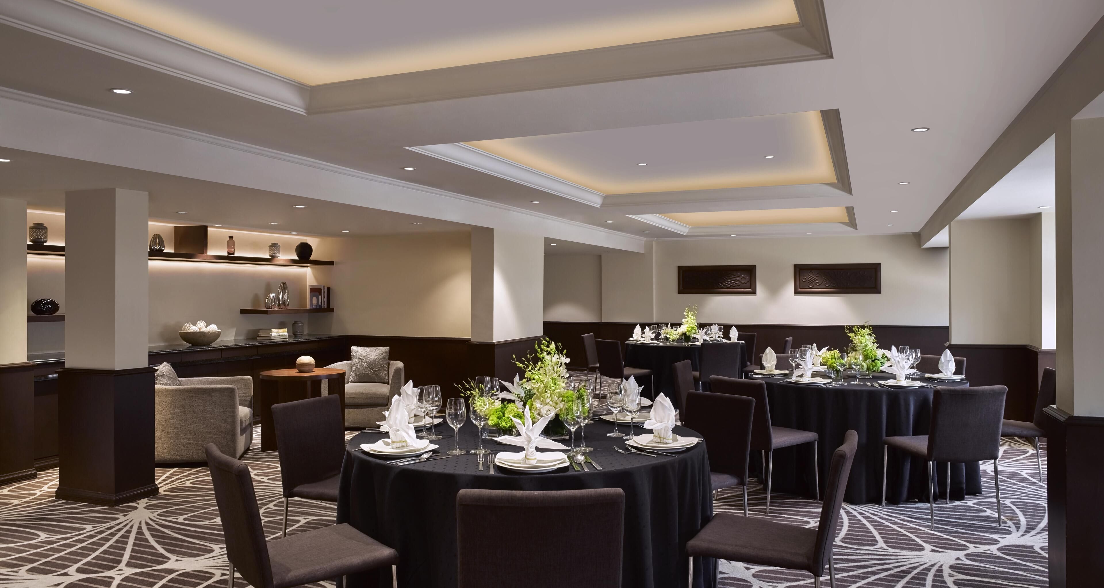 A hotel dining area with round tables set for a meal, featuring dark tablecloths, white plates, and green centerpieces.