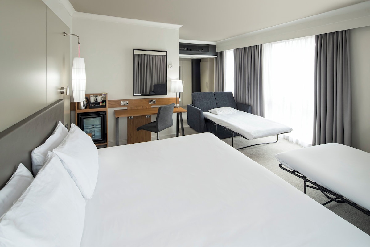 A hotel room with a large bed, a desk with a chair, a small table with a lamp, and a TV.