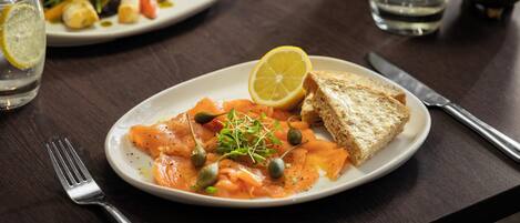 A plate of smoked salmon with capers and a lemon wedge, accompanied by a slice of bread, on a dark wooden table.