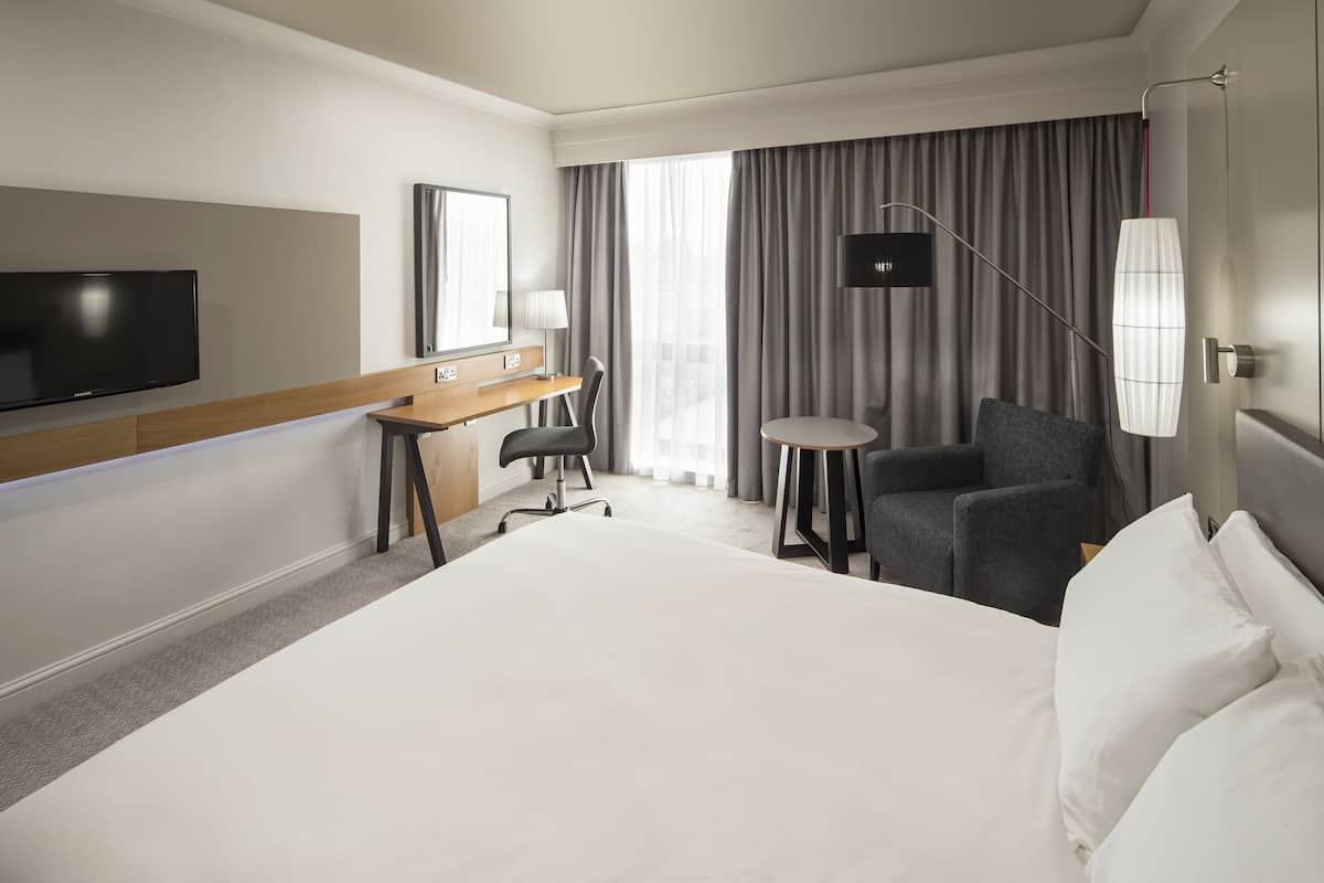 A hotel room with a large bed, a desk with a chair, a small table, a TV, and a lamp.