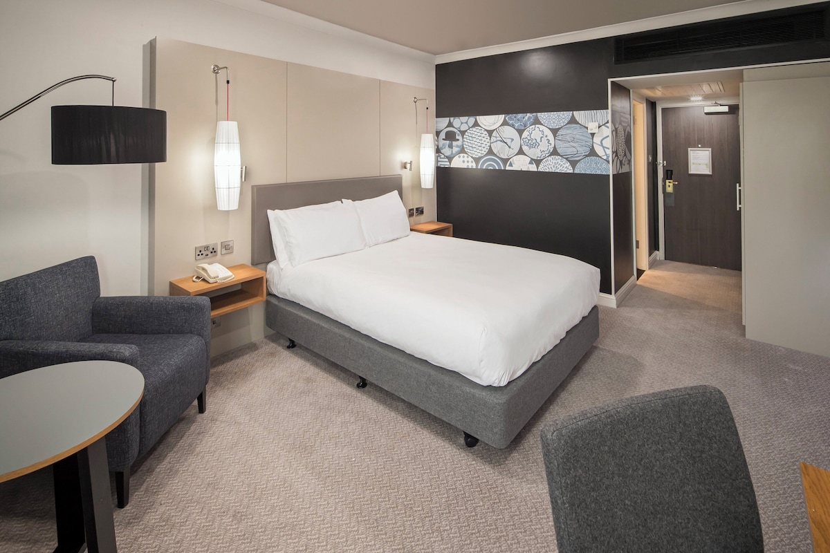 A modern hotel room with a large bed, a desk, a chair, and a small table.