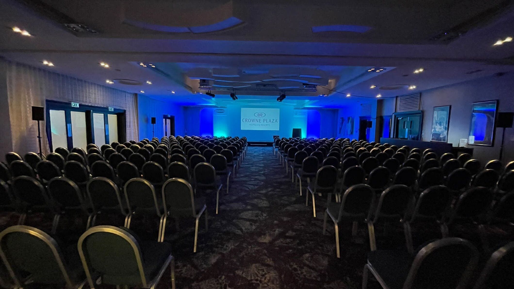 A conference room with rows of chairs facing a presentation screen, a blue-lit ceiling, and a sign reading 'CROWNE PLAZA HOTELS & RESORT'.
