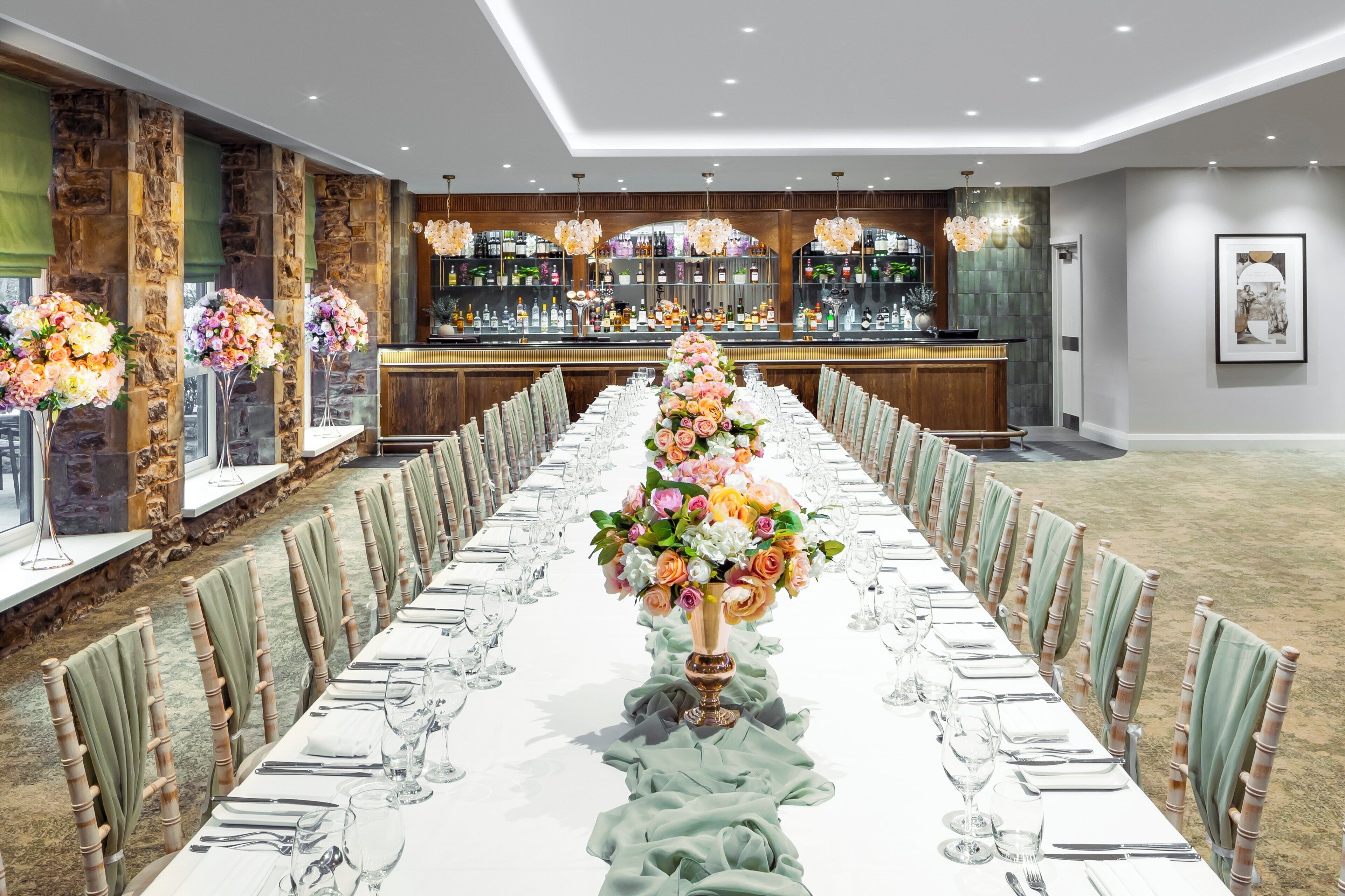 A long banquet table set with floral centerpieces, surrounded by chairs with green cushions.