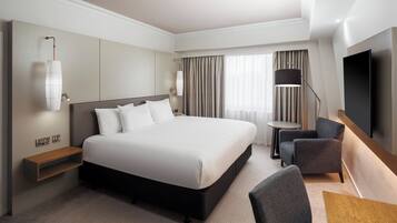 A modern hotel room with a large bed, a desk, a chair, a TV, and a window with curtains.