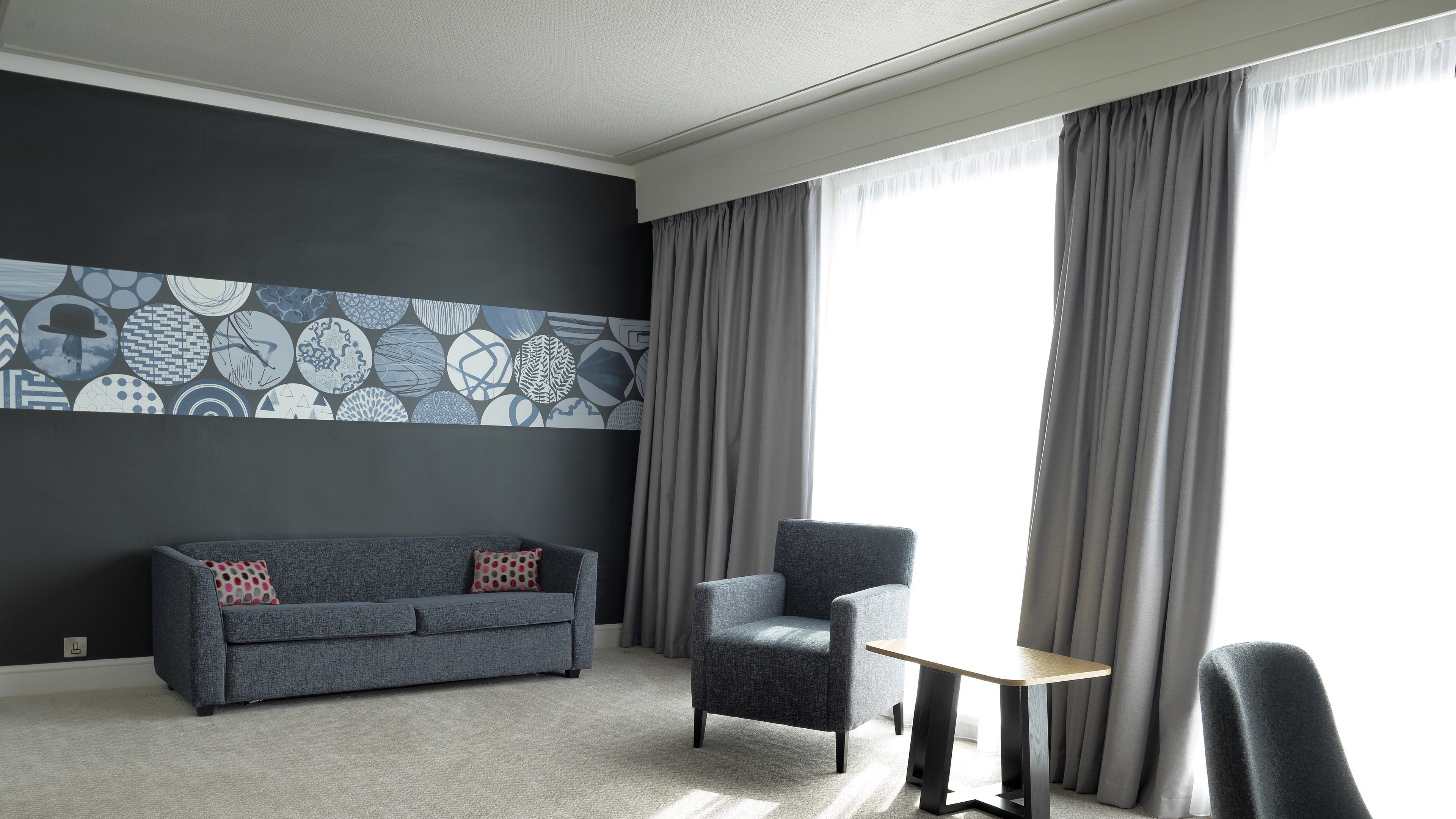 A modern hotel room with a grey sofa, a small table, and a chair. The room features a large window with grey curtains and a wall with a geometric patterned design.