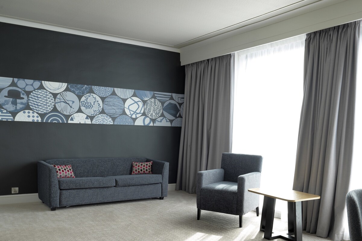 Una habitación de hotel moderna con un sofá gris, una mesita pequeña y una silla. La habitación tiene un ventanal grande con cortinas grises y una pared con un diseño geométrico.