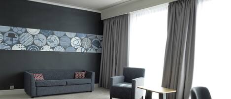 A modern hotel room with a grey sofa, a small table, and a chair. The room features a large window with grey curtains and a wall with a geometric patterned design.