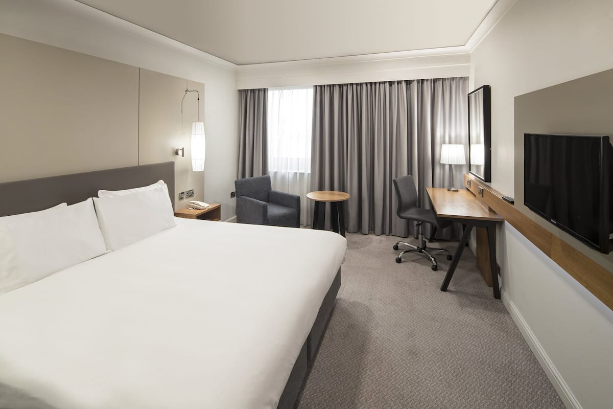 A modern hotel room with a large bed, a desk with a chair, a television, and a large window with curtains.