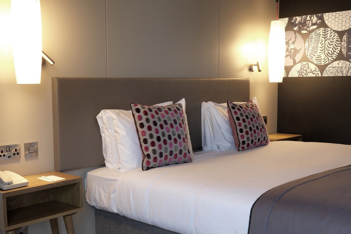 Una habitación de hotel con una cama grande, dos mesitas de noche, un teléfono y lámparas fijadas a la pared.