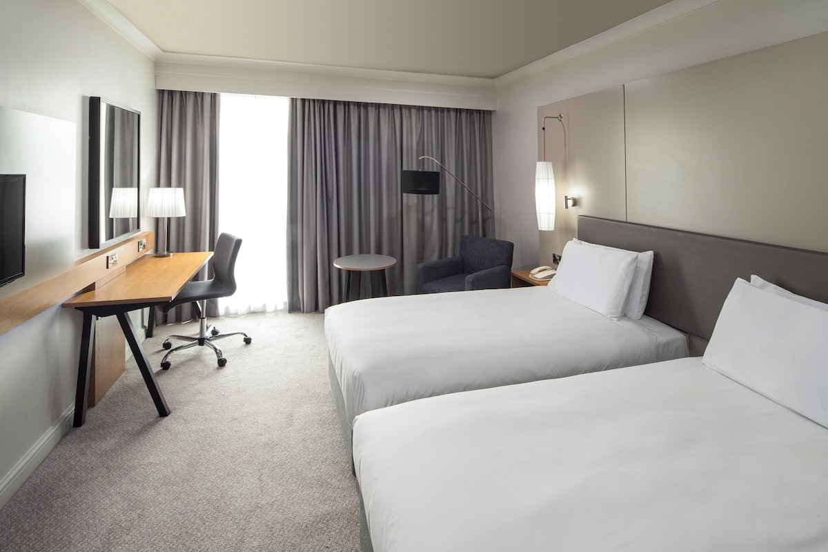 A hotel room with two beds, a desk, a chair, a TV, and a window with curtains.
