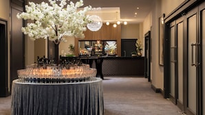 A hotel lobby with a round table covered in a dark tablecloth, surrounded by empty wine glasses, a large white floral arrangement, and a bar area in the background.