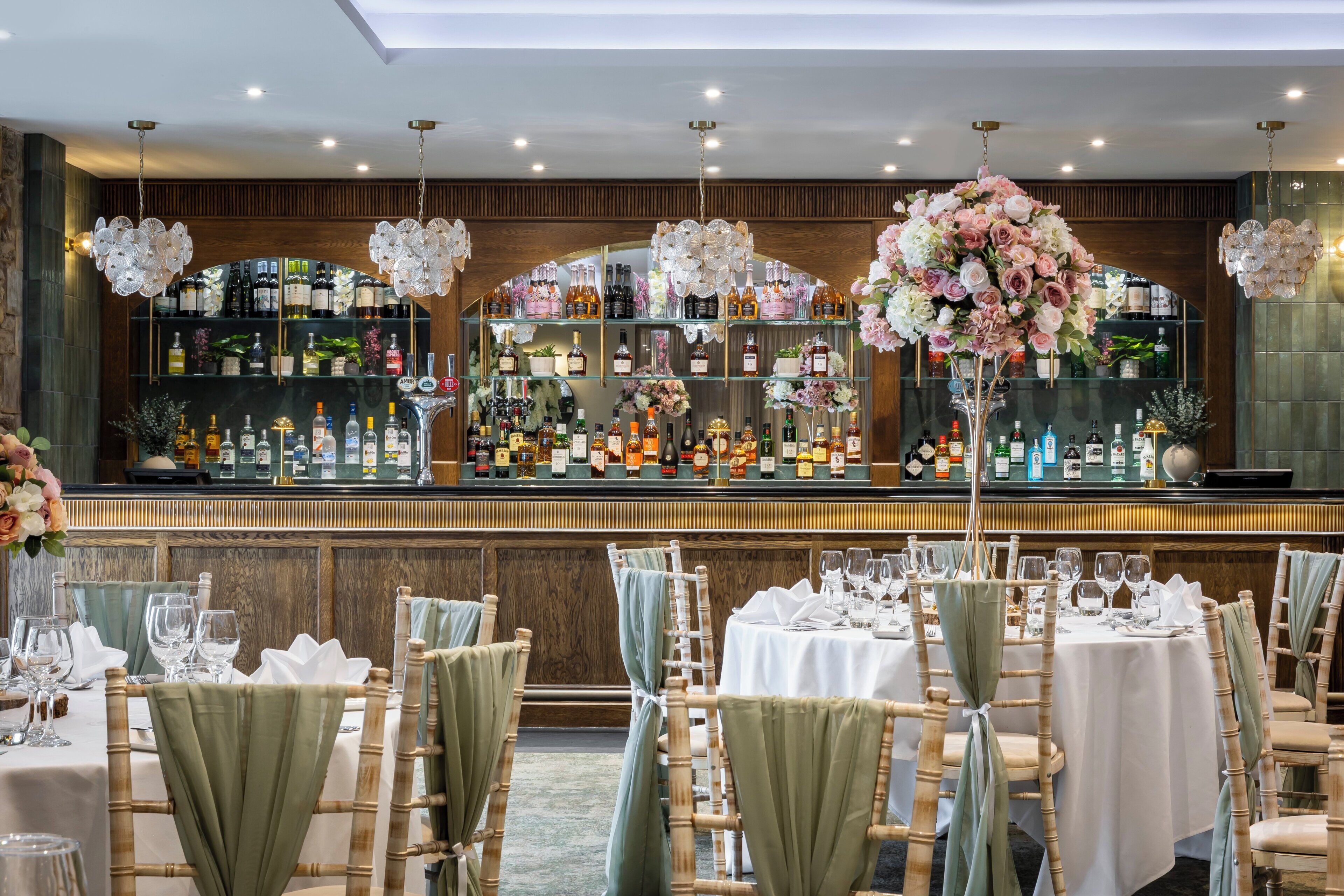 A bar area with neatly arranged tables and chairs, a well-stocked bar with various bottles, and a floral centerpiece.