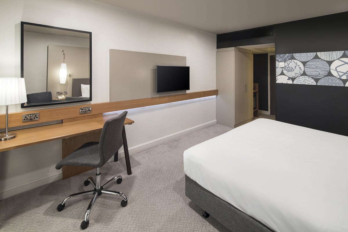 A hotel room with a bed, a desk with a chair, a flat-screen TV, a mirror, and a patterned wall.
