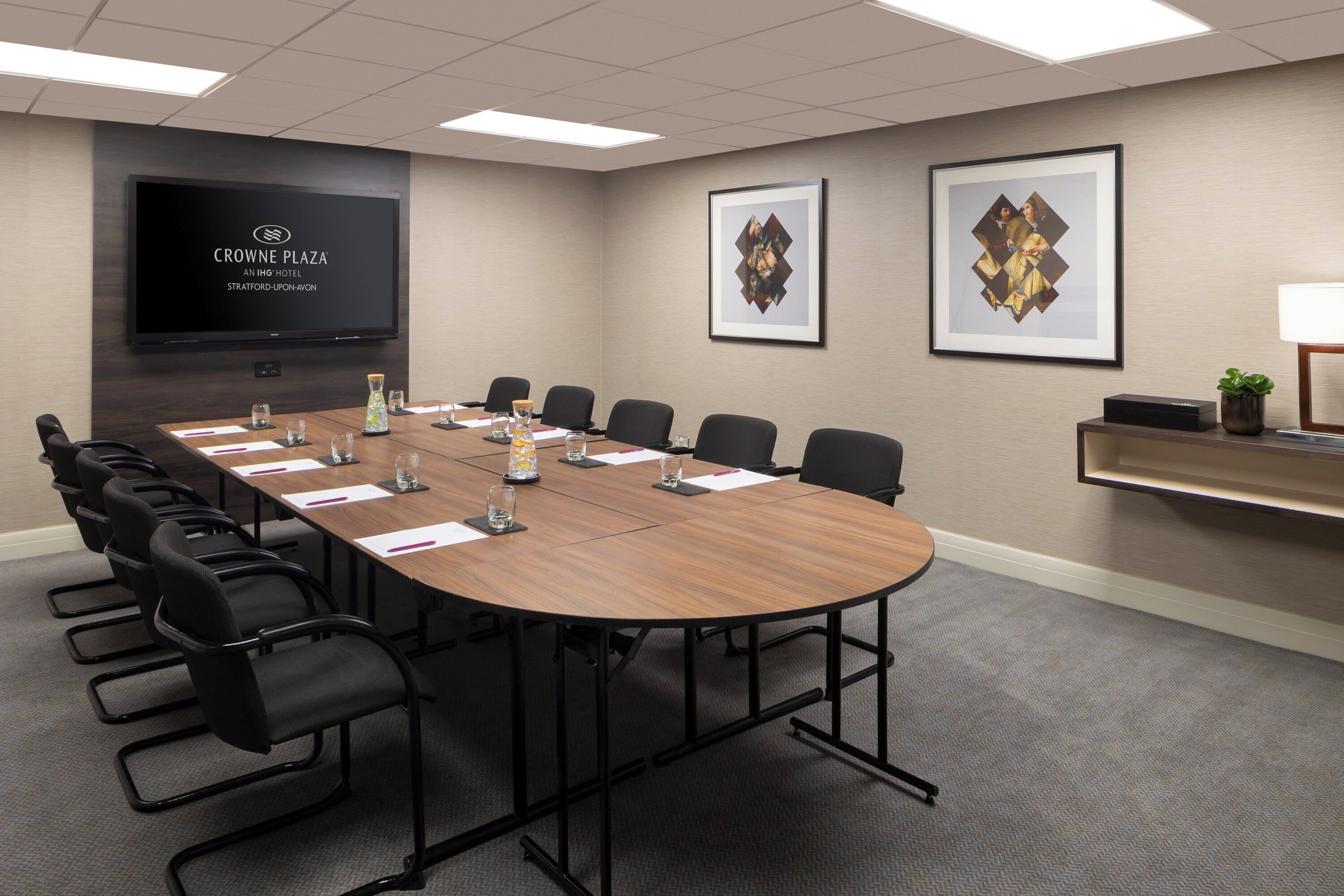 A conference room with a long table, black chairs, a flat-screen TV, framed artwork, and a small plant on a shelf.