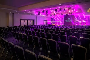 Un salón de conferencias con filas de sillas y un escenario con equipo de iluminación.