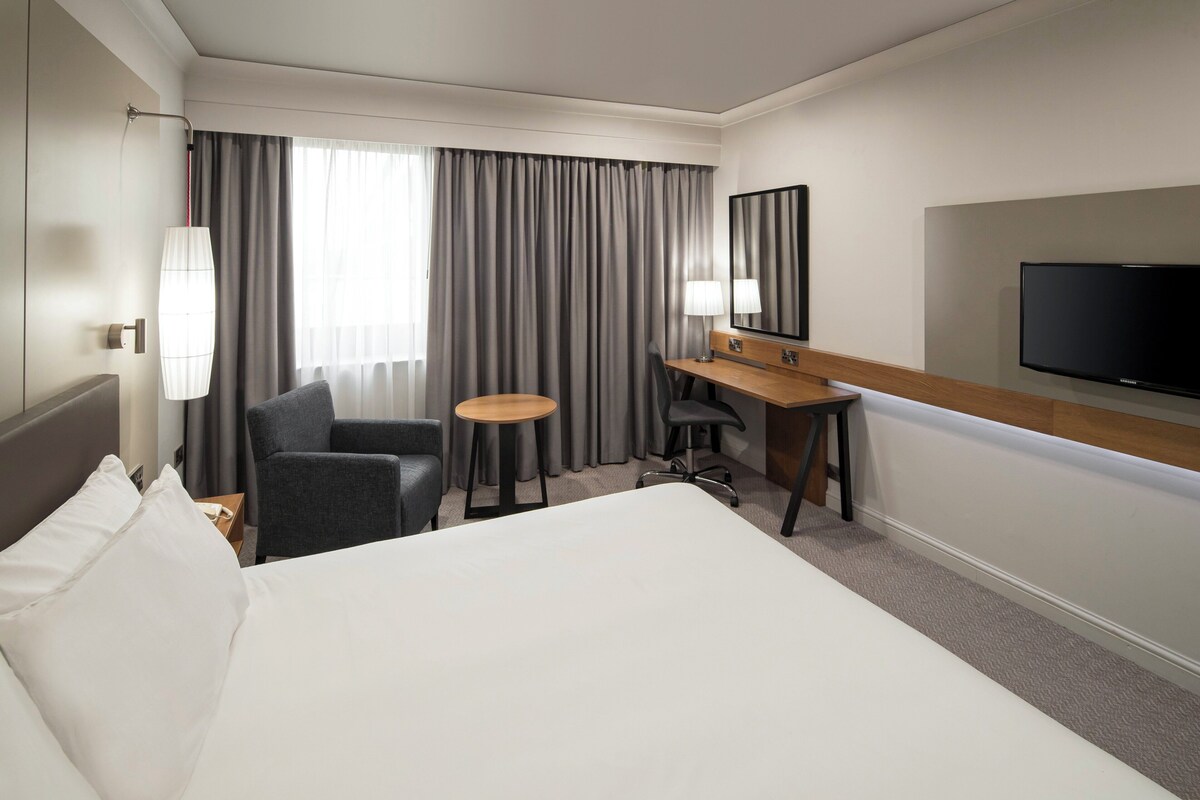 Habitación de hotel con una cama grande, un escritorio, una silla, una mesita, un televisor y una ventana con cortinas.