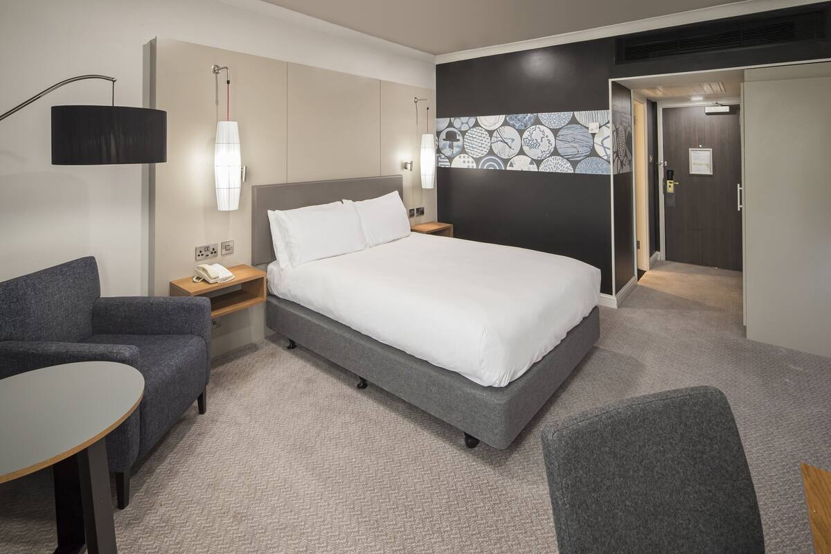 A modern hotel room with a large bed, a desk, a chair, and a small table.