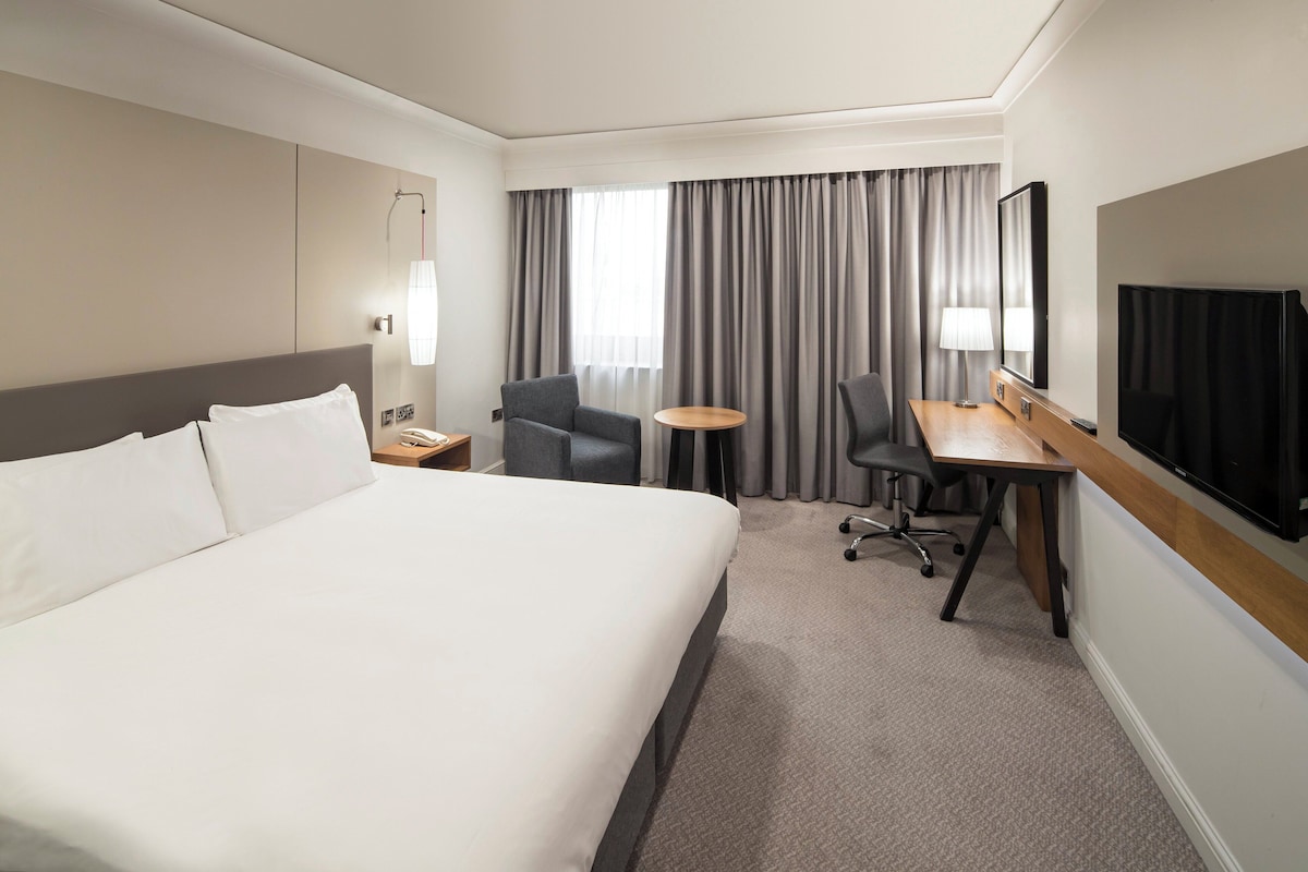 A modern hotel room with a large bed, a desk with a chair, a television, and a large window with curtains.