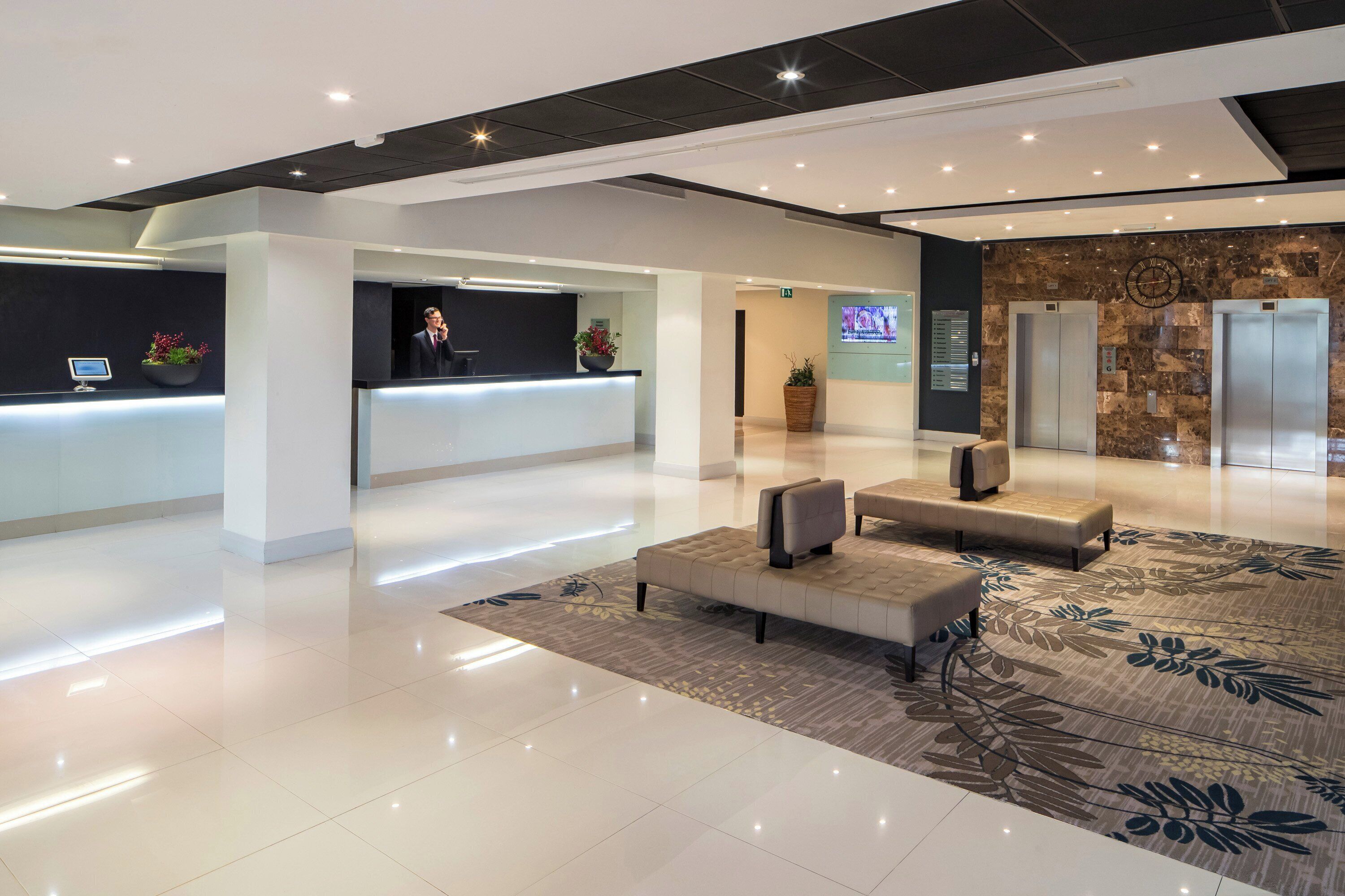Een moderne hotelentree met een receptiebalie, een zithoek, liften en een digitaal scherm.