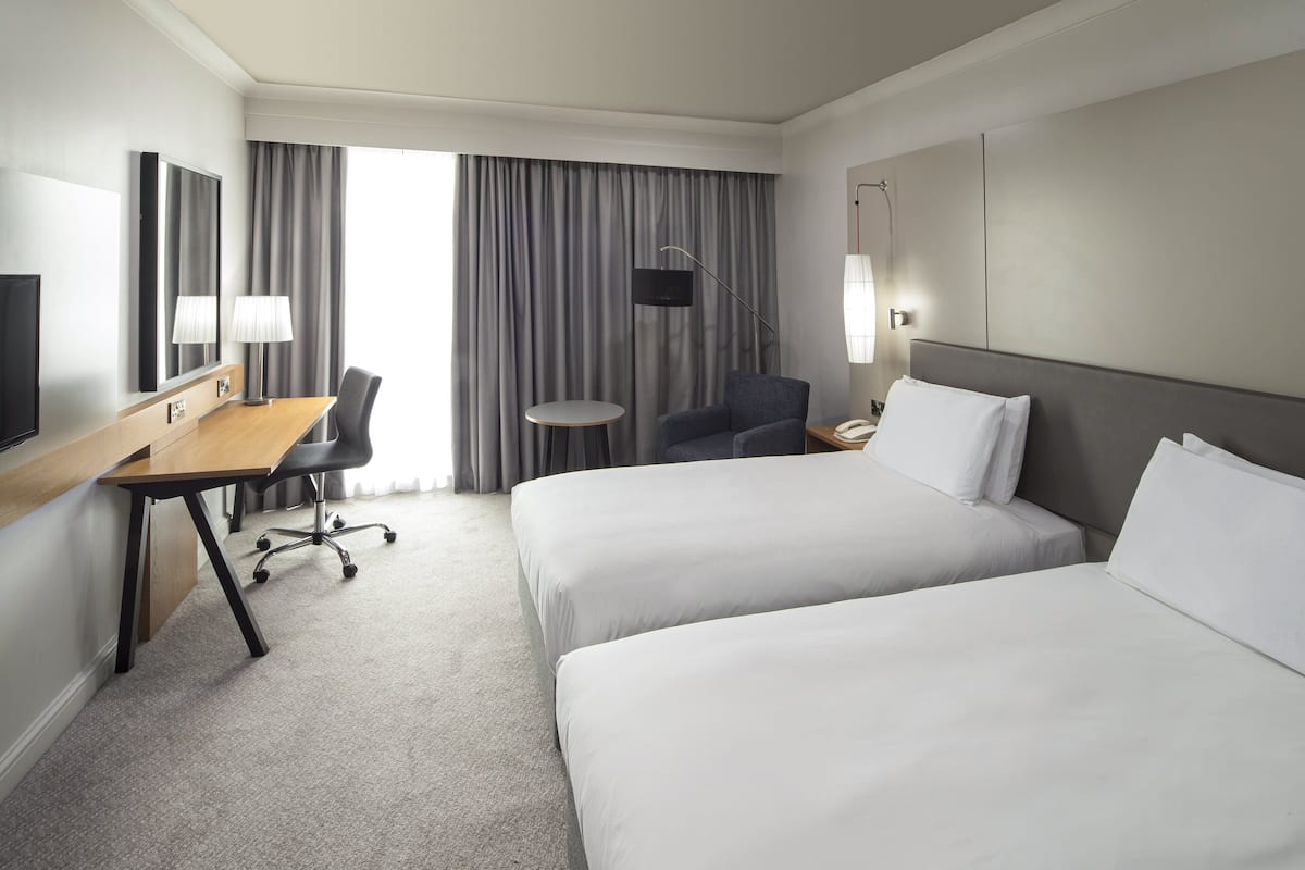 A hotel room with two beds, a desk, a chair, a TV, and a window with curtains.