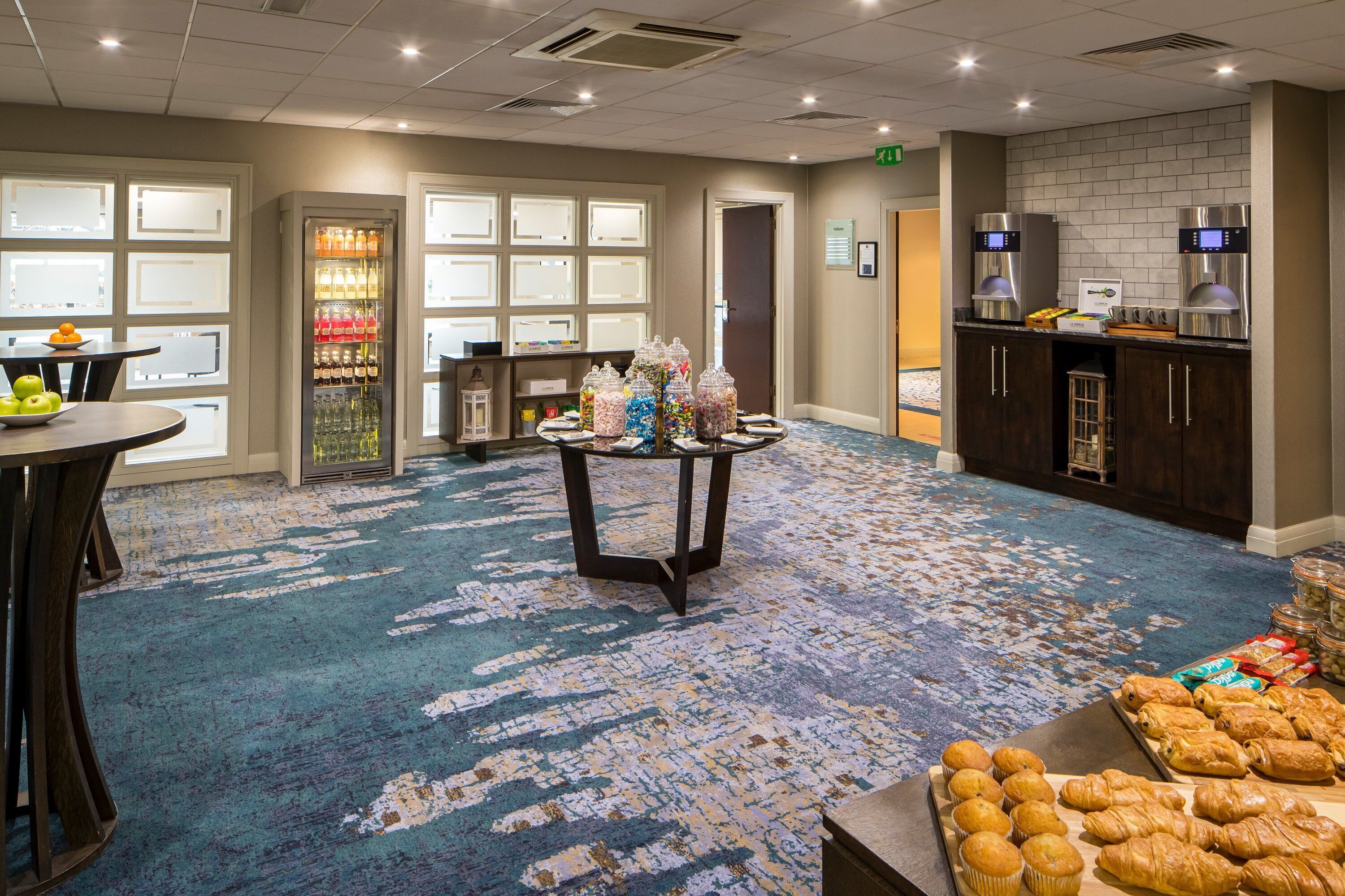 Un hall de hotel con alfombra azul, un frutero, un refrigerador de bebidas y una mesa de buffet con pastelería.