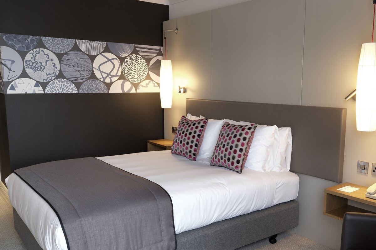 Una habitación de hotel moderna con una cama grande, dos mesitas de noche, un escritorio con teléfono y una pared con diseño geométrico.