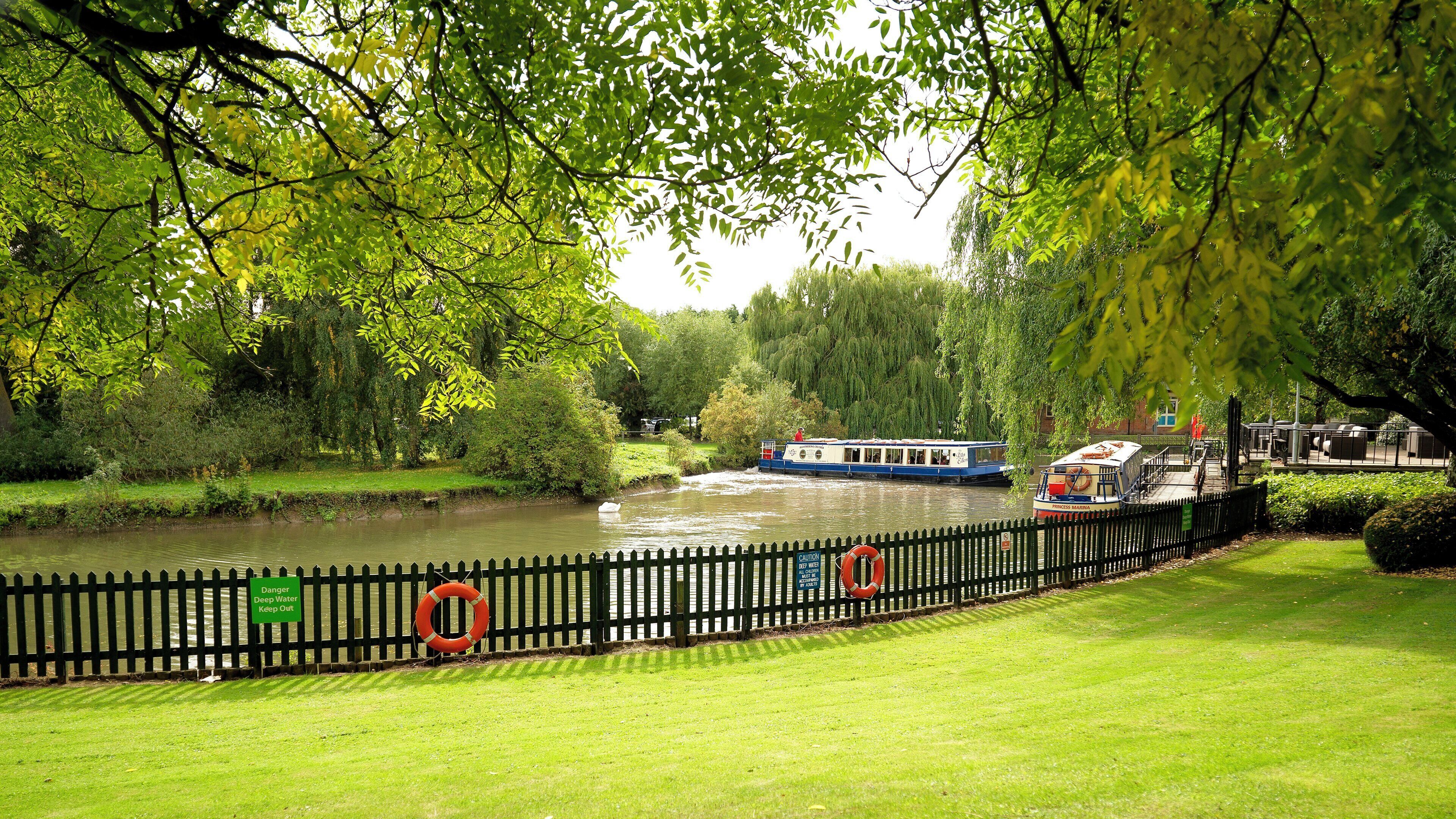 A river with boats, a grassy bank, and a fence with safety signs.