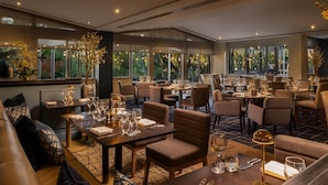 A well-lit restaurant with multiple tables and chairs, large windows with natural light, and decorative plants.
